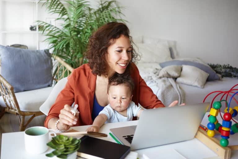 woman working at desk at home using laptop, holding baby on her lap.