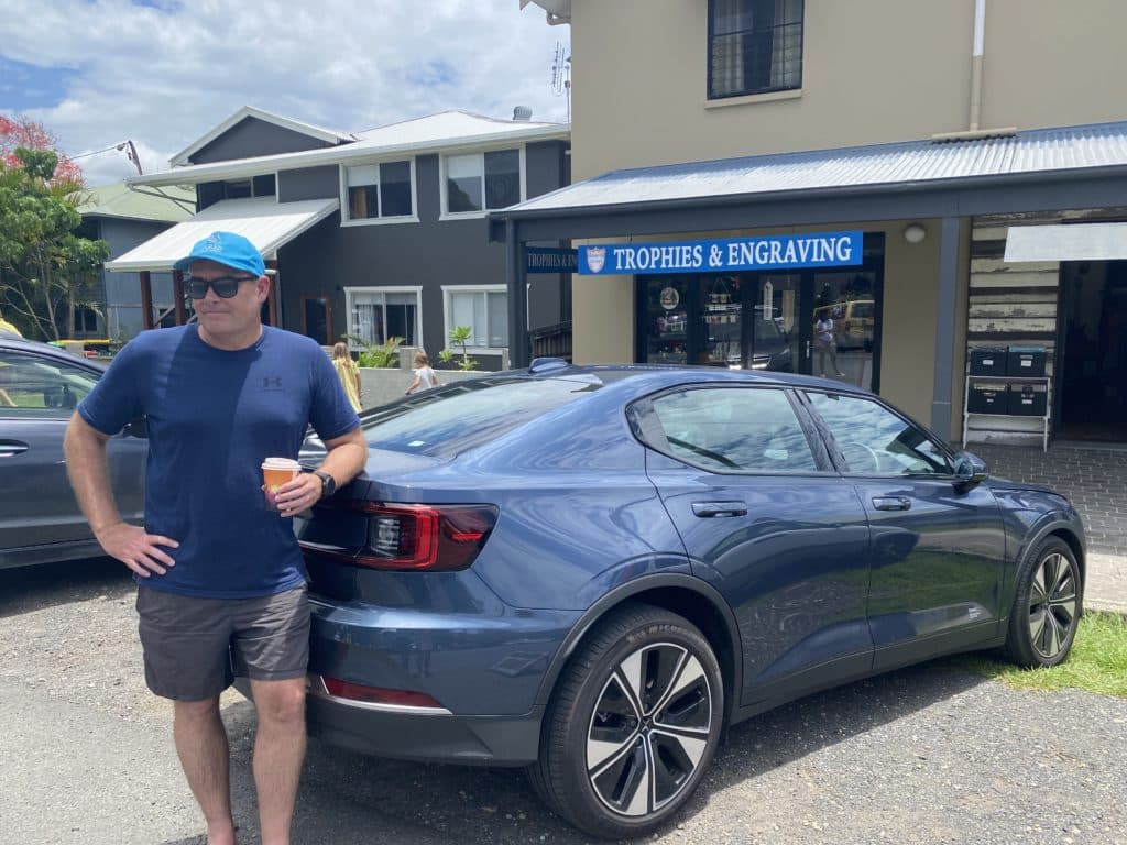 John Horan standing at the rear of a blue sedan wearing a hat